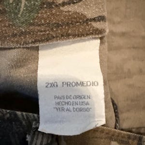 K&ouml;nnte beinhalten: Nahaufnahme eines Camouflage-Stoffs mit einem wei&szlig;en Etikett. Das Etikett lautet "2XG PROMEDIO" und enth&auml;lt spanischen Text: "PAIS DE ORIGEN HECHO EN USA 'VER AL DORSO'". Der Stoff hat gr&uuml;ne, braune und beige Tarnmuster.
