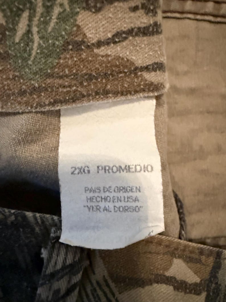 K&ouml;nnte beinhalten: Nahaufnahme eines Camouflage-Stoffs mit einem wei&szlig;en Etikett. Das Etikett lautet "2XG PROMEDIO" und enth&auml;lt spanischen Text: "PAIS DE ORIGEN HECHO EN USA 'VER AL DORSO'". Der Stoff hat gr&uuml;ne, braune und beige Tarnmuster.