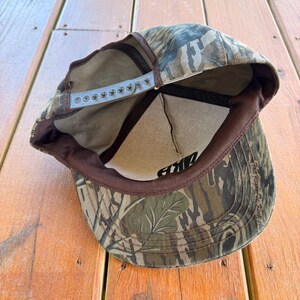 May include: Camouflage baseball cap with a brown brim and adjustable snap closure. The cap features a green, brown, and tan pattern. The interior has a brown band and a white mesh lining. The word "AKE" is visible in black lettering.