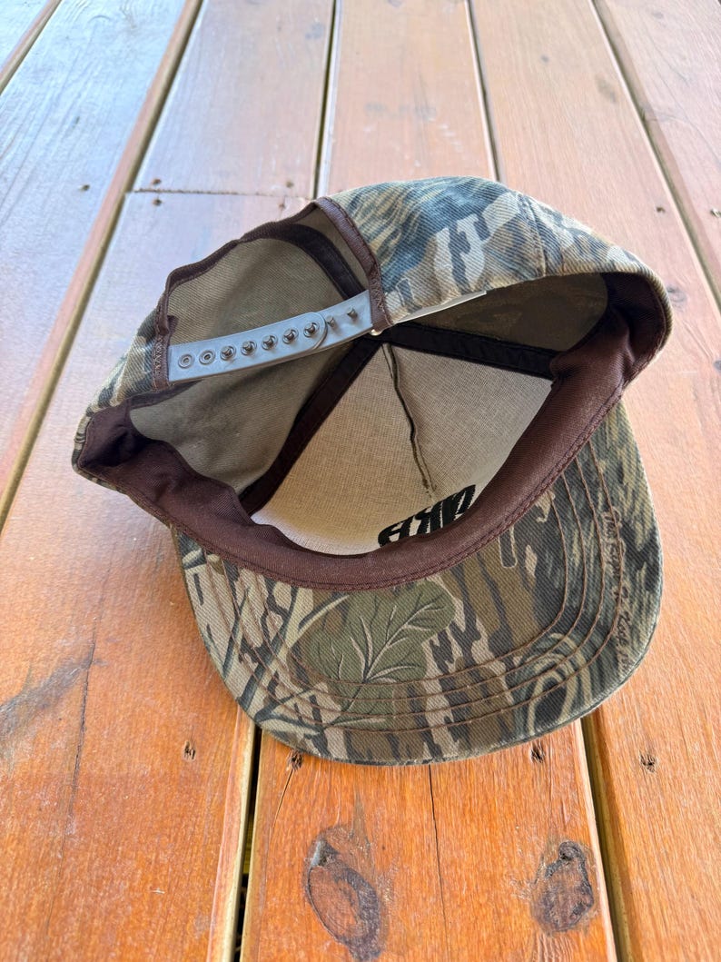 May include: Camouflage baseball cap with a brown brim and adjustable snap closure. The cap features a green, brown, and tan pattern. The interior has a brown band and a white mesh lining. The word "AKE" is visible in black lettering.