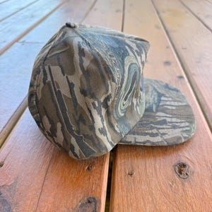 May include: A camouflage baseball cap in shades of brown, green, and tan. The cap has a curved brim and a rounded crown. The fabric has a textured appearance, and the cap is resting on a wooden surface.