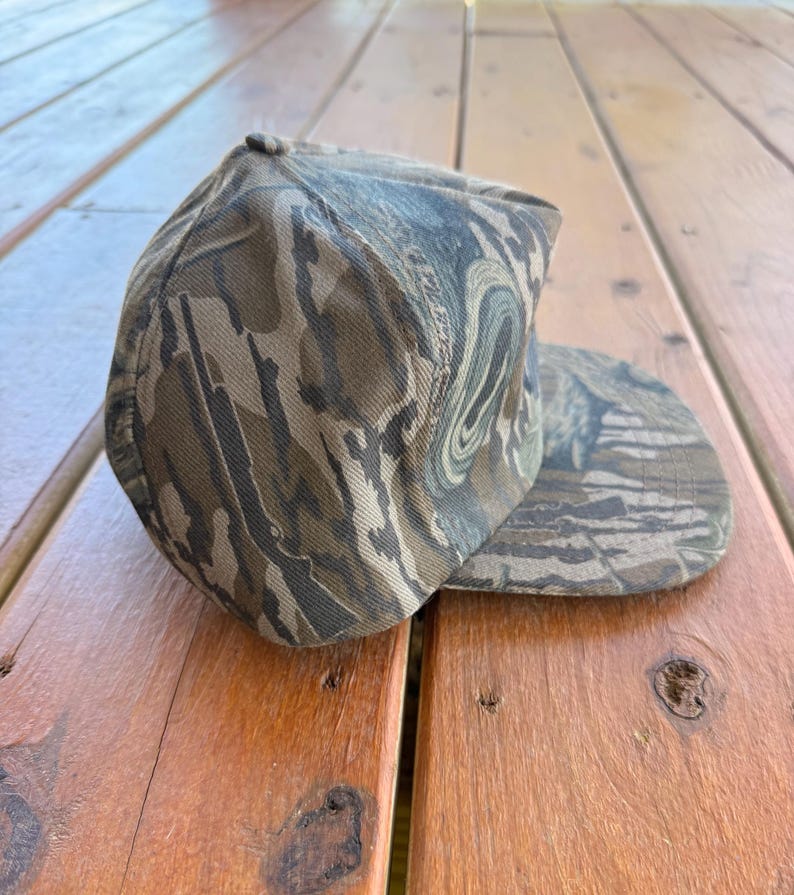 May include: A camouflage baseball cap in shades of brown, green, and tan. The cap has a curved brim and a rounded crown. The fabric has a textured appearance, and the cap is resting on a wooden surface.