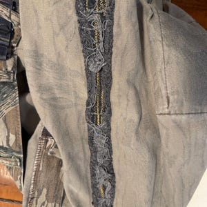 K&ouml;nnte beinhalten: Nahaufnahme eines Kleidungsst&uuml;cks mit Camouflagemuster und grauem Stoff. Ein vertikaler, dekorativer Aufn&auml;her aus grauem Faden mit goldfarbenen N&auml;hten ist auf das Kleidungsst&uuml;ck gen&auml;ht.