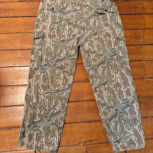 K&ouml;nnte beinhalten: Tarnfarbene Cargohose in einem braunen, gr&uuml;nen und beigen Muster. Die Hose hat mehrere Taschen und einen Knopfverschluss. Das Tarnmuster zeigt ein Astmuster. Die Hose liegt flach auf einer Holzoberfl&auml;che.