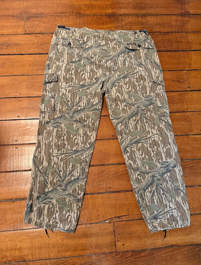 K&ouml;nnte beinhalten: Tarnfarbene Cargohose in einem braunen, gr&uuml;nen und beigen Muster. Die Hose hat mehrere Taschen und einen Knopfverschluss. Das Tarnmuster zeigt ein Astmuster. Die Hose liegt flach auf einer Holzoberfl&auml;che.