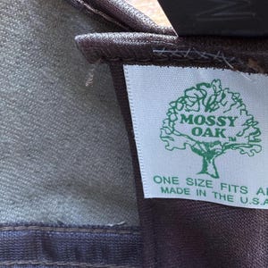 May include: Close-up of a fabric item with a tag. The tag features the "Mossy Oak" logo, a tree graphic, and the text "ONE SIZE FITS ALL MADE IN THE U.S.A." The fabric appears to be a blend of green and brown.