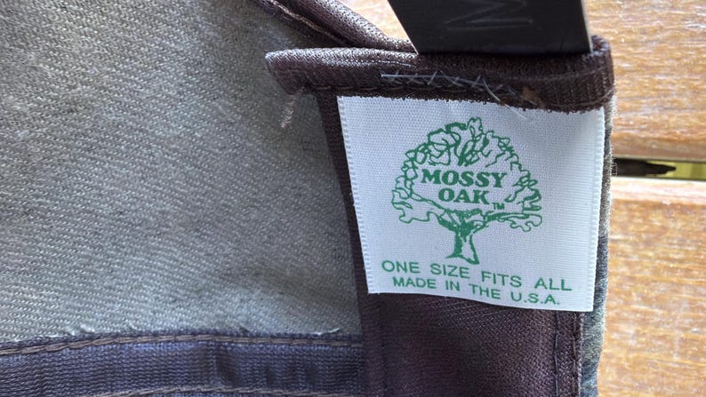 May include: Close-up of a fabric item with a tag. The tag features the "Mossy Oak" logo, a tree graphic, and the text "ONE SIZE FITS ALL MADE IN THE U.S.A." The fabric appears to be a blend of green and brown.