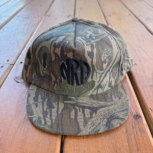 May include: A camouflage baseball cap with a curved brim. The cap features a dark, circular monogram of the letters "NRD" on the front. The hat is made of a fabric with a pattern of brown, green, and tan.