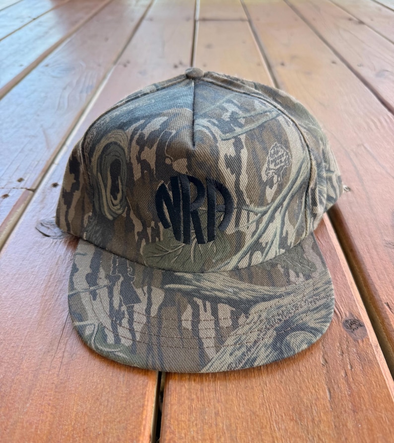 May include: A camouflage baseball cap with a curved brim. The cap features a dark, circular monogram of the letters "NRD" on the front. The hat is made of a fabric with a pattern of brown, green, and tan.
