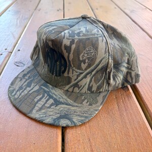 May include: A camouflage baseball cap with a flat brim. The cap features a woodland pattern in shades of brown, green, and tan. The word "MR" is embroidered on the front in black. The cap is made of a durable fabric.