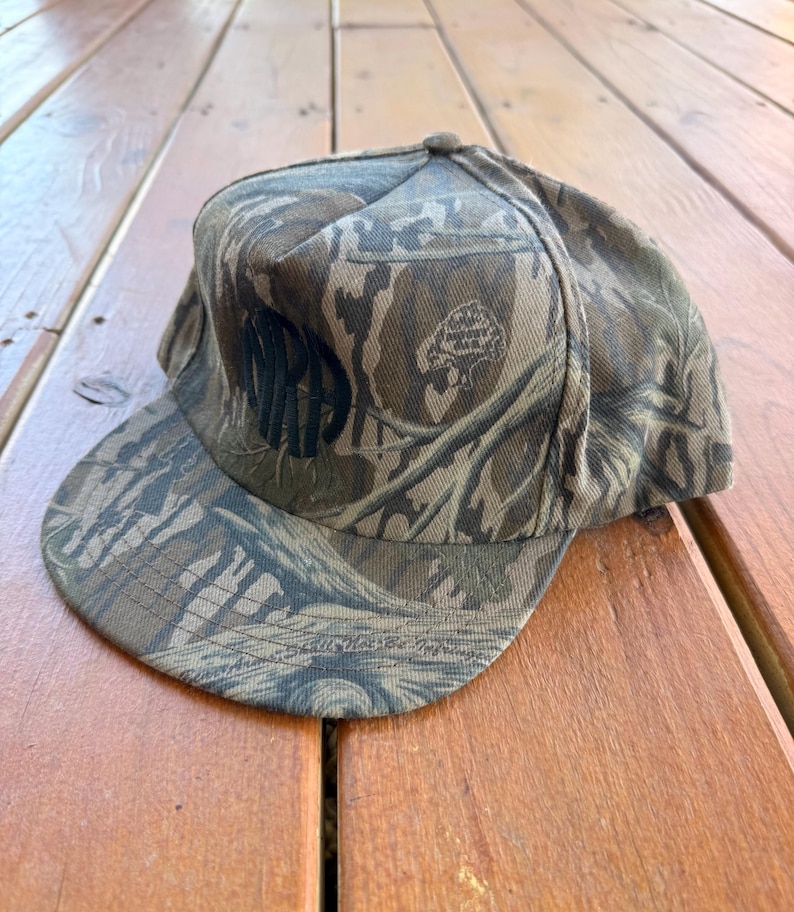 May include: A camouflage baseball cap with a flat brim. The cap features a woodland pattern in shades of brown, green, and tan. The word "MR" is embroidered on the front in black. The cap is made of a durable fabric.