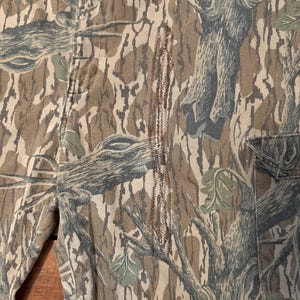 K&ouml;nnte beinhalten: Nahaufnahme einer Camouflage-Hose mit einem Baumrindenmuster in Braun-, Grau- und Gr&uuml;nt&ouml;nen. Die Hose hat sichtbare N&auml;hte und ein Taschen Detail. Der Stoff hat ein strukturiertes Aussehen.