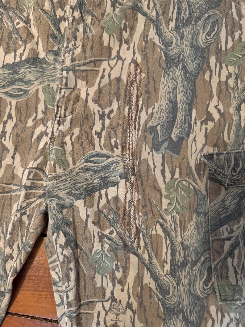 K&ouml;nnte beinhalten: Nahaufnahme einer Camouflage-Hose mit einem Baumrindenmuster in Braun-, Grau- und Gr&uuml;nt&ouml;nen. Die Hose hat sichtbare N&auml;hte und ein Taschen Detail. Der Stoff hat ein strukturiertes Aussehen.