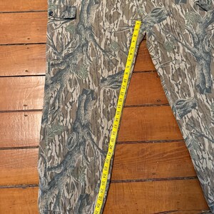 K&ouml;nnte beinhalten: Tarnfarbene Cargohose mit einem Waldmuster in Braun-, Gr&uuml;n- und Graut&ouml;nen. Die Hose hat mehrere Taschen und wird mit einem gelben Ma&szlig;band gemessen. Das Ma&szlig; betr&auml;gt etwa 84 cm.