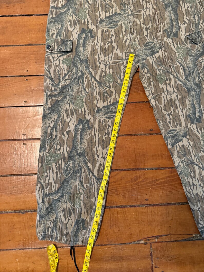 K&ouml;nnte beinhalten: Tarnfarbene Cargohose mit einem Waldmuster in Braun-, Gr&uuml;n- und Graut&ouml;nen. Die Hose hat mehrere Taschen und wird mit einem gelben Ma&szlig;band gemessen. Das Ma&szlig; betr&auml;gt etwa 84 cm.