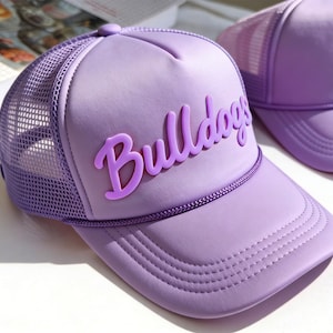 Puede incluir: Dos gorras de camionero color lavanda. La parte delantera de la gorra presenta la palabra "Bulldogs" en una fuente cursiva en relieve. La gorra tiene una parte trasera de malla y una visera curva. Las gorras están sobre una superficie blanca.