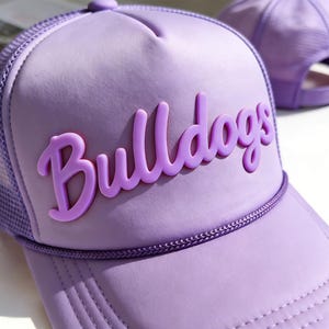 Puede incluir: Gorra de camionero lavanda con la palabra "Bulldogs" en letras en relieve en la parte delantera. La gorra tiene una parte trasera de malla y una visera curva. Una segunda gorra es visible en el fondo.