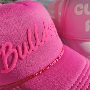 May include: Close-up of two pink trucker hats. The front hat has the word "Bulldog" in raised lettering. The second hat, partially visible, has the word "Custom" in raised lettering. Both hats have a mesh back and a rope detail.