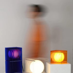 May include: Three modern lamps with a geometric design. One lamp is blue with a purple glow, another is clear with a white light, and the third is orange with a yellow glow. Each lamp has a rectangular cage-like structure.