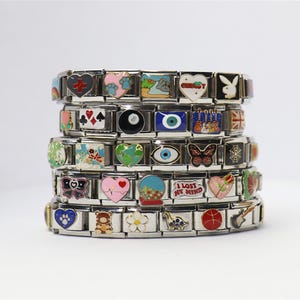 May include: Stack of silver charm bracelets with various colorful charms. Charms include hearts, butterflies, and other designs. The bracelets are stacked on top of each other against a white background.
