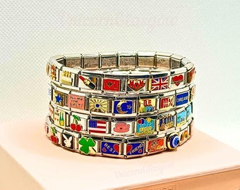 Op de afbeelding: Stapel zilveren bedelarmbanden met kleurrijke emaille bedels. Bedels bevatten vlaggen, harten en teksten als "You are the best!" en "Happy Birthday". De armbanden liggen op een roze doos.