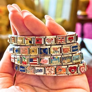 May include: Several charm bracelets are held in a hand. The bracelets are silver-toned with rectangular charms. Each charm features colorful designs, including hearts, flags, and text like "LOVE", "MOM", and "Happy Birthday".