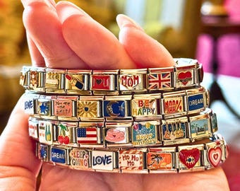 Op de afbeelding: Een hand houdt meerdere bedelarmbanden vast. De zilverkleurige armbanden hebben rechthoekige bedels. Elke bedel heeft kleurrijke ontwerpen, waaronder harten, vlaggen en teksten als "LOVE", "MOM" en "Happy Birthday".