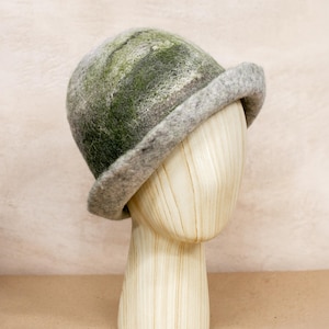 Puede incluir: Un sombrero de fieltro con una copa redondeada y un ala vuelta hacia arriba. El sombrero presenta un patrón veteado de tonos verdes y grises. Se exhibe sobre una cabeza de maniquí de madera, mostrando su forma y diseño. El sombrero es un accesorio elegante.