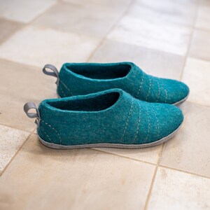May include: A pair of teal blue felt slippers with a light grey trim. The slippers have a simple design with stitching details.