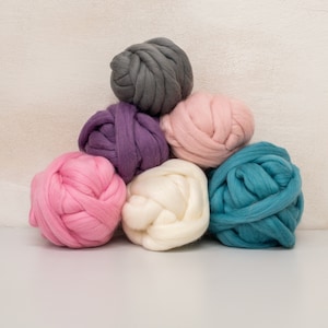 May include: A pile of six colorful yarn balls in shades of pink, purple, gray, white, and teal. The yarn is thick and fluffy, perfect for knitting or crocheting.