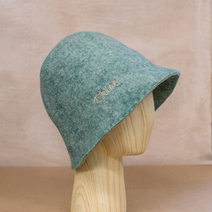 May include: A teal felt bucket hat with a rounded crown and a short, slightly flared brim. The name Chloe is embroidered in a light-coloured script font on the side of the hat. The hat is displayed on a light-coloured wooden mannequin head.