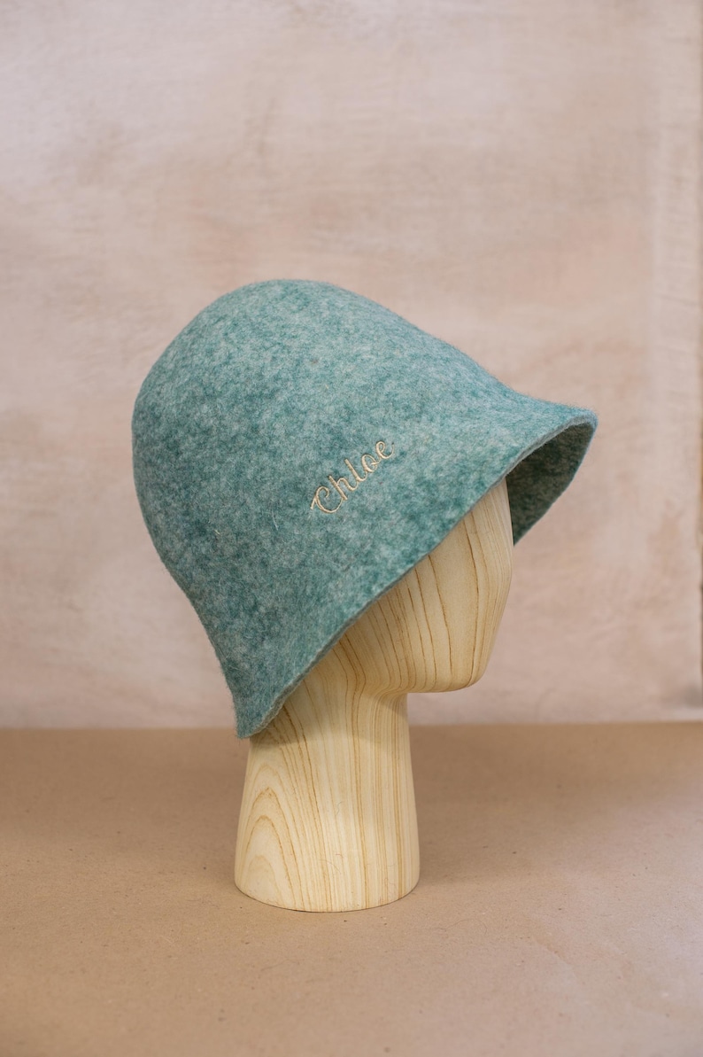 May include: A teal felt bucket hat with a rounded crown and a short, slightly flared brim. The name Chloe is embroidered in a light-coloured script font on the side of the hat. The hat is displayed on a light-coloured wooden mannequin head.