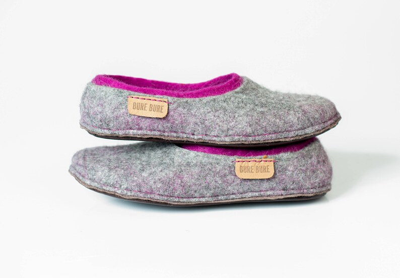K&ouml;nnte beinhalten: Ein Paar graue Filzpantoffeln mit rosa Filzfutter. Die Pantoffeln haben ein braunes Lederlabel mit dem Text "Bure Bure" darauf.