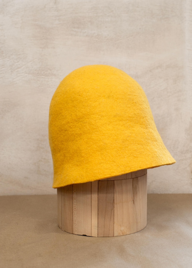 May include: A bright yellow felted wool hat with a rounded crown and a slightly flared brim. The hat features a dense, textured surface characteristic of felted material, showcasing a solid, vibrant colour throughout its entire structure.