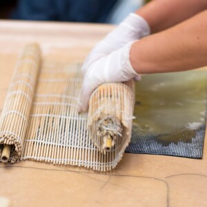 May include: A person is rolling up a bamboo mat used for making sushi. The mat is made of thin strips of bamboo and is held together by string. The mat is being rolled up on a light brown surface.
