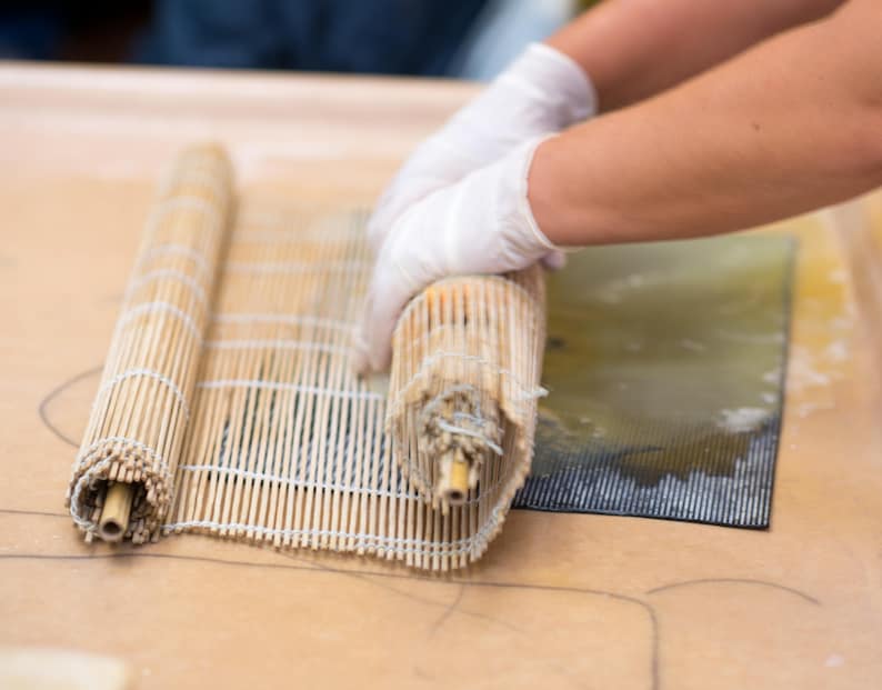 May include: A person is rolling up a bamboo mat used for making sushi. The mat is made of thin strips of bamboo and is held together by string. The mat is being rolled up on a light brown surface.