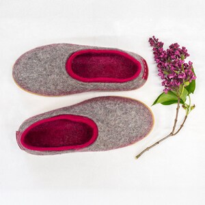 K&ouml;nnte beinhalten: Ein Paar graue Filzpantoffeln mit rosa Filzfutter. Die Pantoffeln liegen auf einem wei&szlig;en Hintergrund mit einem Zweig lila Bl&uuml;ten.