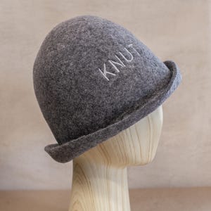 May include: A charcoal grey felted hat with a rounded crown and a slightly upturned brim. The word KNUT is embroidered in white thread on the front of the hat. The hat is displayed on a light-coloured wooden mannequin head form.