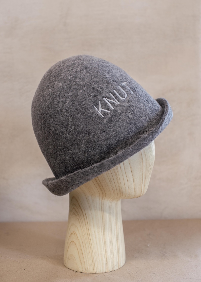 May include: A charcoal grey felted hat with a rounded crown and a slightly upturned brim. The word KNUT is embroidered in white thread on the front of the hat. The hat is displayed on a light-coloured wooden mannequin head form.