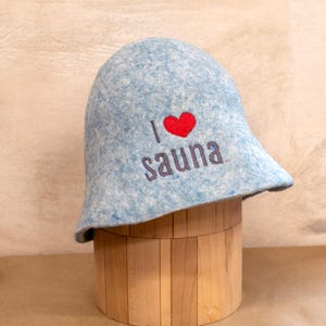 May include: A light blue felted sauna hat with a mottled texture. The front features embroidered text that reads I, followed by a red heart symbol, and the word sauna in dark grey lettering. The hat has a rounded crown and a flared brim.