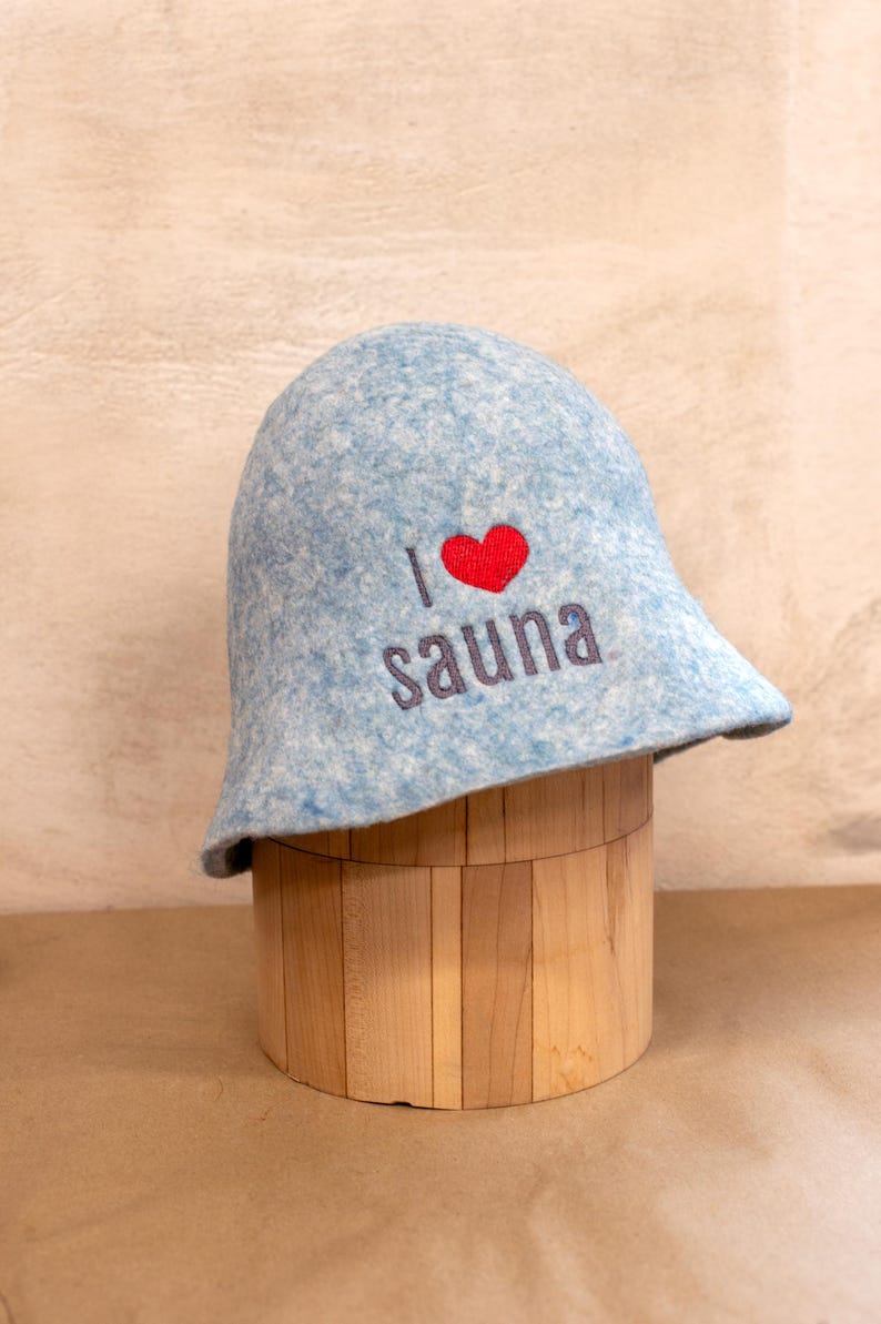 May include: A light blue felted sauna hat with a mottled texture. The front features embroidered text that reads I, followed by a red heart symbol, and the word sauna in dark grey lettering. The hat has a rounded crown and a flared brim.