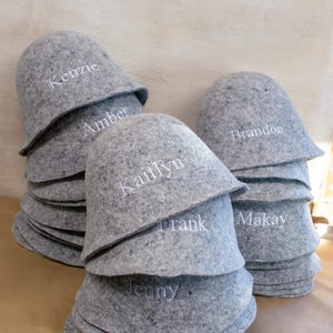 May include: A stack of grey felt sauna hats, each featuring white embroidered names including Kenzie, Amber, Kaitlyn, Frank, Jenny, Brandon, and Makay. The hats are rounded with a wide brim, constructed from a textured, mottled grey material.