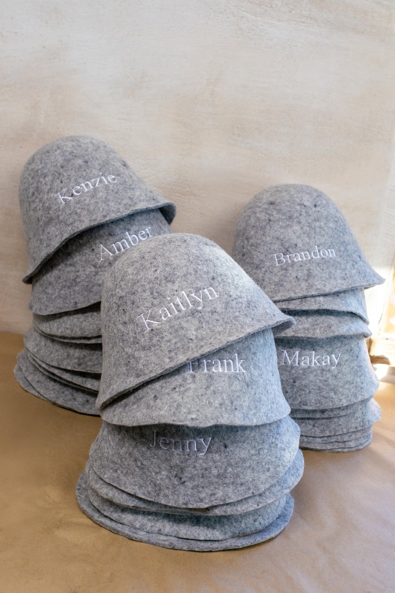 May include: A stack of grey felt sauna hats, each featuring white embroidered names including Kenzie, Amber, Kaitlyn, Frank, Jenny, Brandon, and Makay. The hats are rounded with a wide brim, constructed from a textured, mottled grey material.