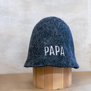 May include: A dark grey felted wool sauna hat with a rounded crown and a wide brim. The word PAPA is embroidered in white capital letters on the front of the hat. The hat has a textured, fuzzy surface and a structured, conical shape.