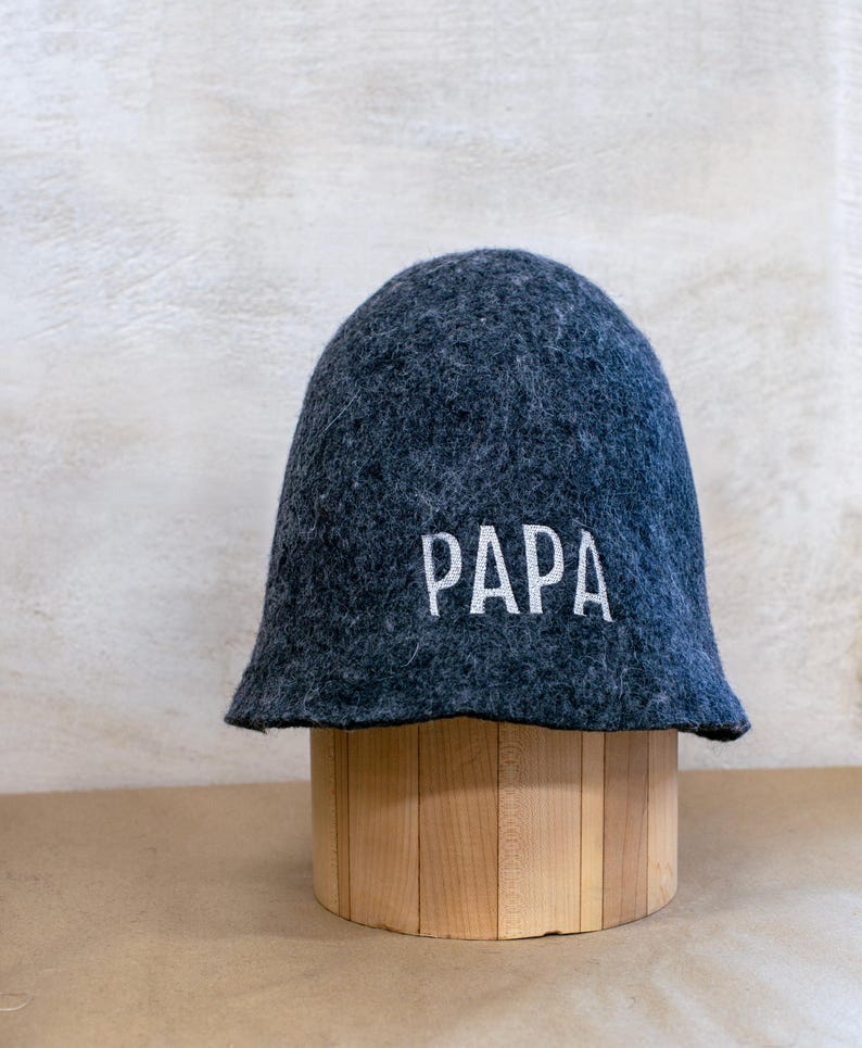 May include: A dark grey felted wool sauna hat with a rounded crown and a wide brim. The word PAPA is embroidered in white capital letters on the front of the hat. The hat has a textured, fuzzy surface and a structured, conical shape.