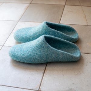 May include: A pair of light blue felted wool slippers. The slippers are a solid color and have a soft, textured appearance. The open-back design provides easy wear. The slippers are placed on a tiled surface.