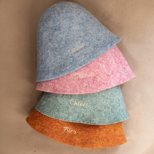 May include: A stack of four felted wool bucket hats in blue, pink, teal, and orange. Each hat features a personalised name embroidered in white thread on the front: Timma on the blue hat, Gabriel on the pink, Chloe on the teal, and Alex on the orange.
