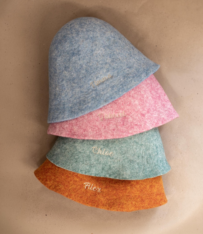 May include: A stack of four felted wool bucket hats in blue, pink, teal, and orange. Each hat features a personalised name embroidered in white thread on the front: Timma on the blue hat, Gabriel on the pink, Chloe on the teal, and Alex on the orange.