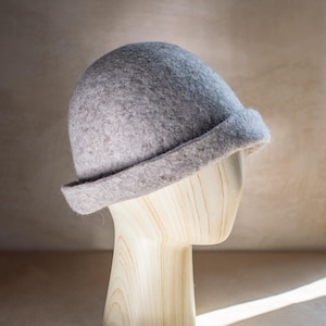 May include: A light grey felted hat with a rounded crown and a short, turned-up brim. The hat features a soft, textured surface and is displayed on a light-coloured wooden mannequin head, showcasing its simple, structured, and classic form.