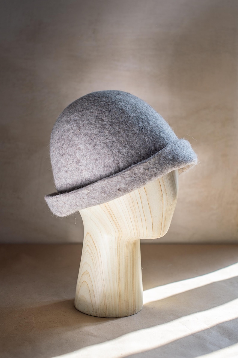 May include: A light grey felted hat with a rounded crown and a short, turned-up brim. The hat features a soft, textured surface and is displayed on a light-coloured wooden mannequin head, showcasing its simple, structured, and classic form.